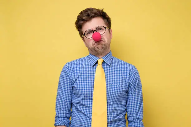 Sad caucasian man wearing clown red nose on yellow wall. He is upset that nobody came to his party.