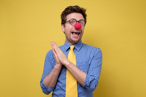 Handsome young man with red clown nose smiling and clapping hands. Studio shot on yellow wall.