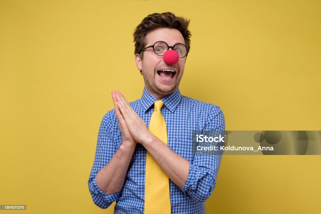 Handsome young man with red clown nose smiling and clapping hands. Handsome young man with red clown nose smiling and clapping hands. Studio shot on yellow wall. Clown Stock Photo