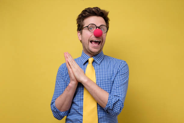 Handsome young man with red clown nose smiling and clapping hands. Handsome young man with red clown nose smiling and clapping hands. Studio shot on yellow wall. fool stock pictures, royalty-free photos & images