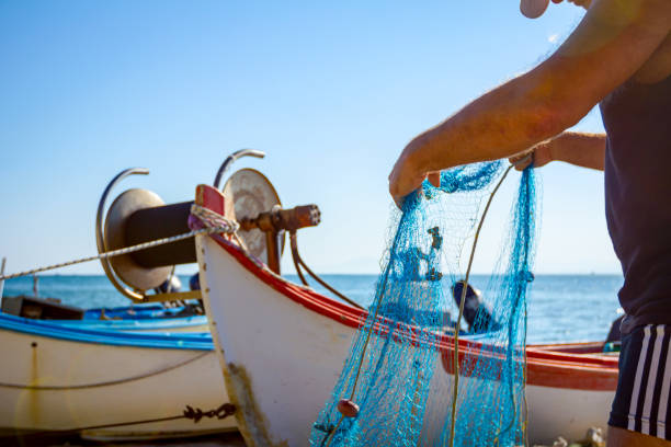 Makrygialos, Greece - August 23, 2018: Fisherman pile up fishnet and prepare for his next angling. Boats are dry docked on the sandy beach.