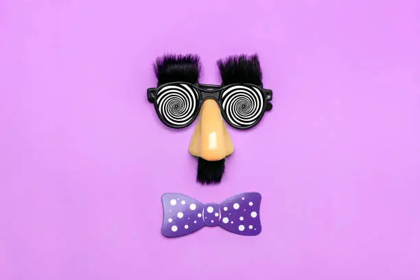 Photo of funny face - fake eyeglasses, nose and mustache, confetti, sequins on purple background Happy fools day concept 1st April party Holiday card