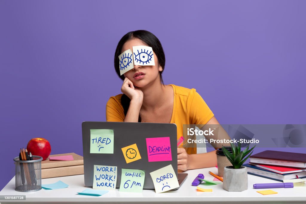 Tired indian woman sitting at desk with sticky notes A Lot Of Work Concept. Portrait of tired female freelancer sleeping at workplace with stickers on eyes, sitting at table over purple studio background. Distracted woman at home office Emotional Stress Stock Photo