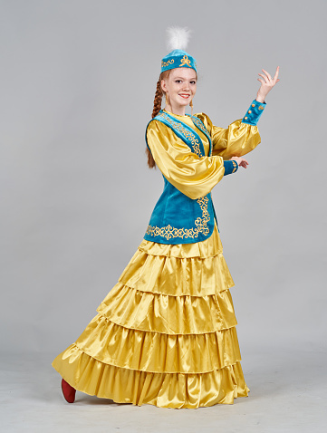 A young caucasian woman with redhead braided hair is dressed in a Kazakh traditional clothing. The young female dancer is  showing traditional poses. She is smiling looking at the camera. Studio shooting