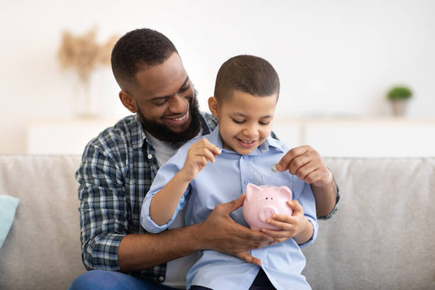African Father And Son Putting Money In Piggybank At Home Financial Literacy. Cheerful African Father And Son Putting Money In Piggybank Sitting On Sofa At Home. Daddy Teaching His Child Budget Planning, Keeping Personal Savings Safety literacy photos stock pictures, royalty-free photos & images