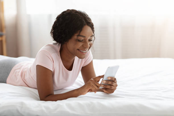 istock Happy black lady laying on bed, using mobile phone 1301591170