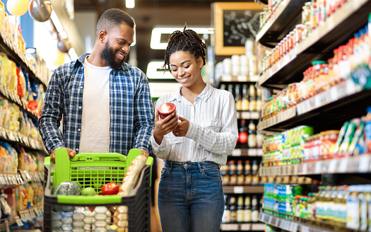 African Family Buying Food In Supermarket Shop Walking Pushing Cart And Choosing Groceries Together. Happy Customers. Black Couple In Grocery Store. Empty Space For Text