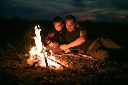 Father and little cute son sitting by the fire in the forest. Best friends spending time together.