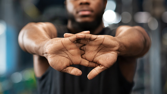Hands in lock of unrecognizable black guy sportsman stretching hands and fingers in gym, closeup, panorama. Cropped of african american young man bodybuilder warming up before exercising