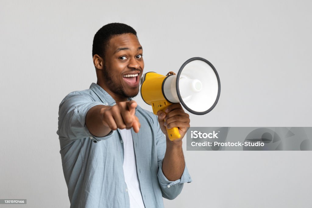 Positive african guy shouting in megaphone over grey background Positive african american guy shouting in megaphone and pointing at camera over grey background, copy space. Happy black man screaming with loudspeaker, cheering up, making advertisement People Stock Photo