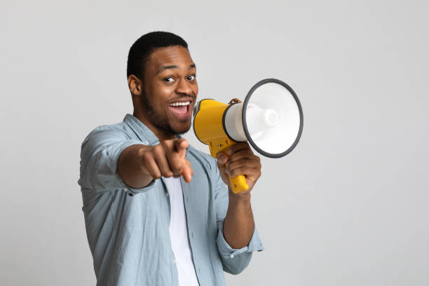 Positive african guy shouting in megaphone over grey background Positive african american guy shouting in megaphone and pointing at camera over grey background, copy space. Happy black man screaming with loudspeaker, cheering up, making advertisement pointing stock pictures, royalty-free photos & images