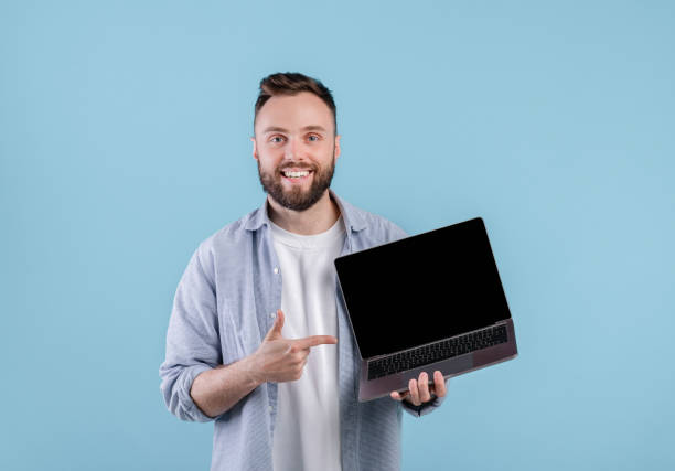 istock Positive young man pointing at empty laptop screen with space for your website design over blue background, mockup 1301596639