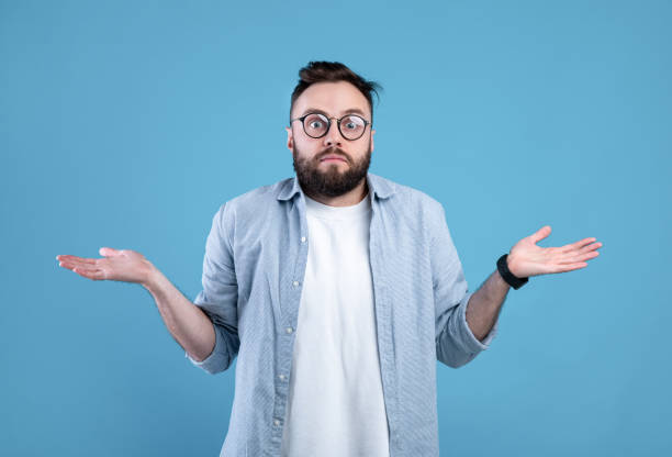 istock Who knows. Confused bearded man in glasses shrugging shoulders on blue studio background 1301596733