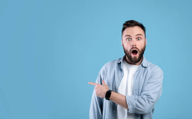 istock Shocked young guy standing with open mouth, pointing at empty space in disbelief, blue background, banner design 1301596799