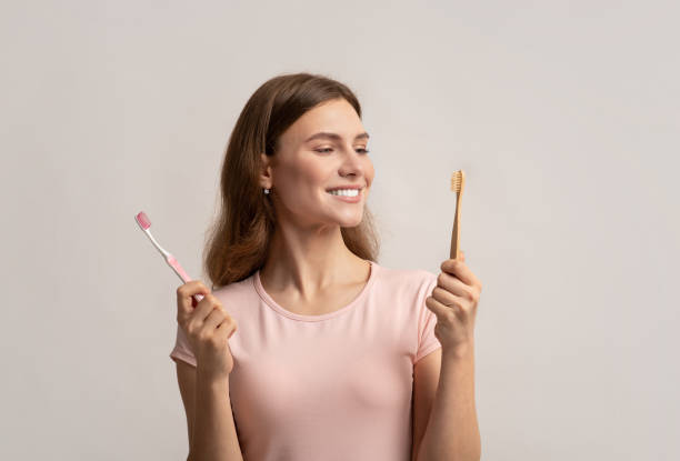 Smiling young woman holding eco bamboo and plastic toothbrush Smiling young woman holding eco bamboo and plastic toothbrush, choosing between different items for dental hygiene. Eco-friendly female looking at wooden brush, standing on grey background, copy space toothbrush stock pictures, royalty-free photos & images
