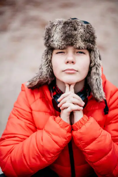 November 21, 2020 - Warsaw, Poland: Handsome intelligent teen boy sitting, looking at camera, hands on chin, looking serious, confident, challenging, smart, intense, wearing fur Russian hat, red jacket - close up, portrait, headshot.
