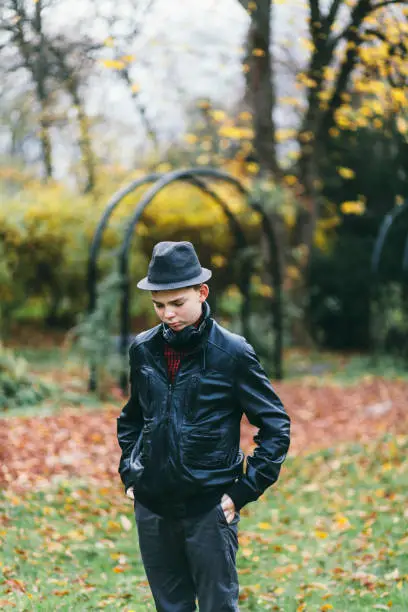 November 21, 2020 - Warsaw, Poland: sad, serious teen boy standing, waiting, hands in pockets, in a park, wearing leather jacket, fedora hat, headphones, looking down, pensive.