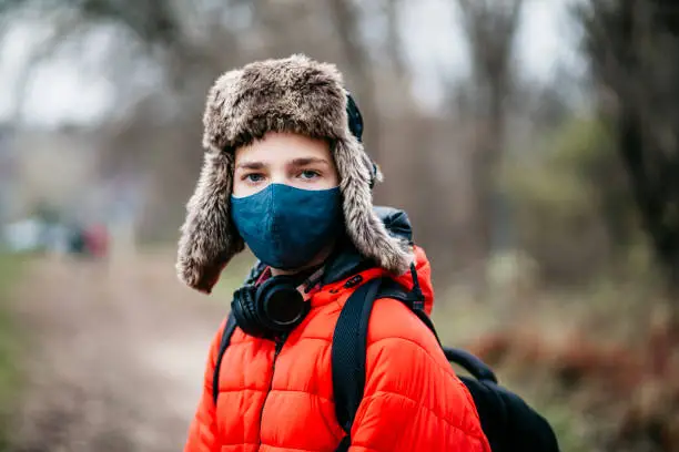 November 21, 2020 - Warsaw, Poland: Headshot of handsome teen boy standing in a park, wearing protective antivirus mask, red jacket, fur Russian hat, headphones, backpack, looking at camera, being sad, worried, interested, curious, tired, bored. Youth culture and coronavirus portrait.