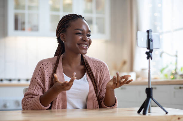 Black Female Blogger Recording Video At Home With Smartphone On Tripod, African American Lady Making Content For Her Blog, Sitting In Kitchen Interior Talking And Gesturing At Phone Camera, Free Space