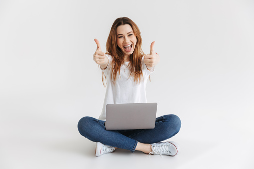 Cheerful woman in t-shirt sitting on the floor with laptop computer while showing thumbs up and looking at the camera over grey background