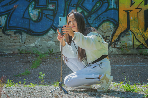 A young girl in a white tracksuit squatted down to set up her mobile phone to record a video of the history of a photo session against the background of a wall with a pattern