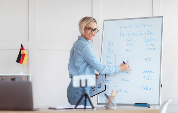 istock Cheerful foreign languages tutor teaching German online using smartphone and laptop, writing on blackboard 1301600151