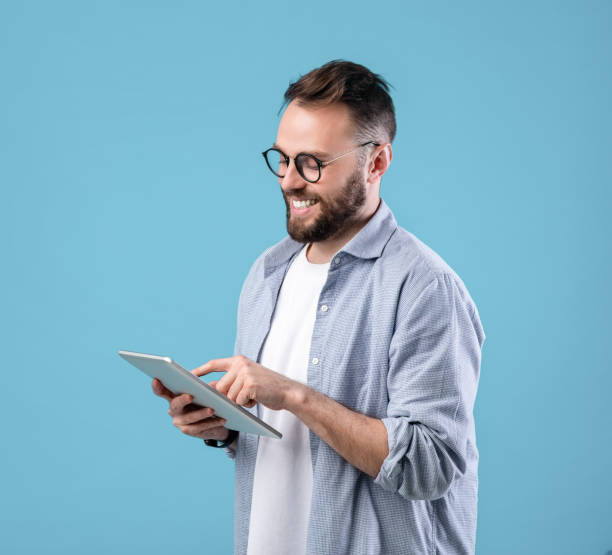 istock Positive young man in glasses using digital tablet for online work or studies on blue studio background 1301601531