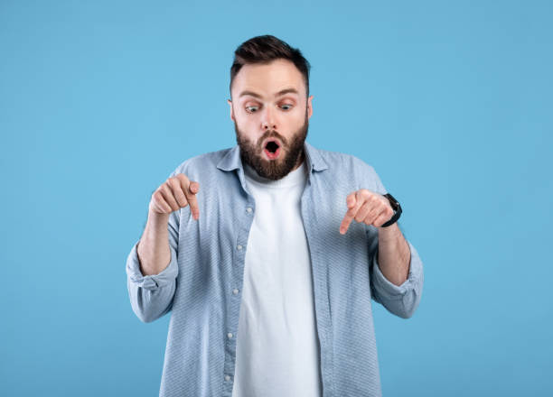 Shocked young bearded man pointing downward at blank space over blue studio background, free space