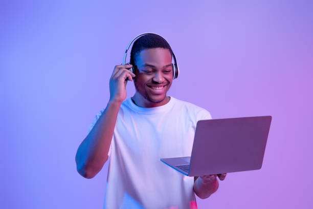 Black Gamer Man In Headset Playing Video Games On Laptop, Neon Lighting Black Gamer Man In Wireless Headset Playing Video Games On Laptop, Looking At Computer Screen While Standing In Vivid Neon Light Over Purple Studio Background, Guy Enjoying Cyber Gaming, Copy Space headset photos stock pictures, royalty-free photos & images