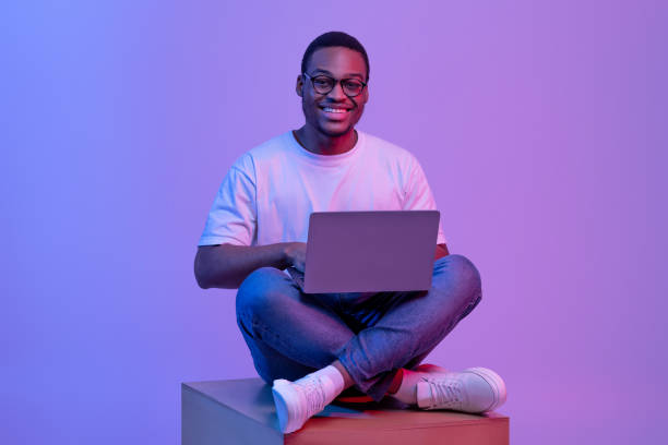 Happy Black Student Guy In Eyeglasses Sitting With Laptop In Neon Lighting Happy Black Student Guy In Eyeglasses Sitting With Laptop On Big Cube In Neon Lighting, Smiling At Camera. Positive Young African American Man Study Or Working Online Over Purple Background In Studio fluorescent photos stock pictures, royalty-free photos & images