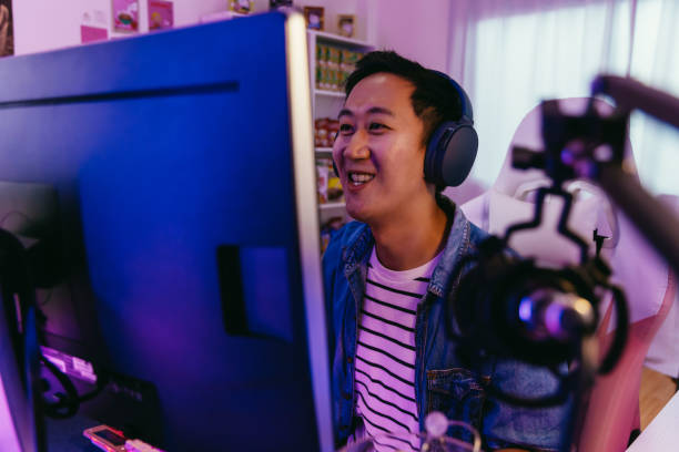 aufgeregt und lächelnde männliche spieler in headset mit mikrofon spielen ein online-videospiel. junger asiatischer mann im gespräch mit spielern und publikum auf pc zu hause - nerd computer learning fun stock-fotos und bilder