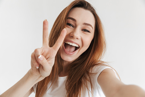 Joyful woman in casual clothes making selfie while showing peace gesture and looking at the camera over grey background