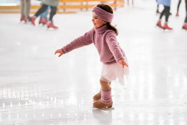 Photo of little girl in pink sweater is skating on a winter evening on an outdoor ice rink lit by garlands