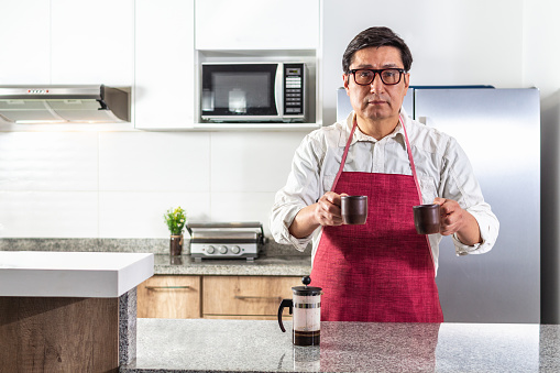 Man in red apron and glasses handing over two cups of French press brewed coffee at a kitchen counter.