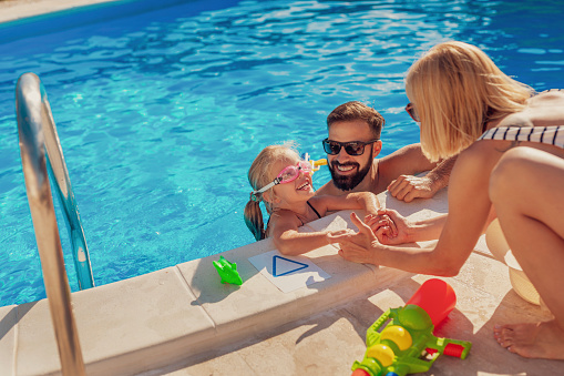 High angle view of young parents having fun playing with their daughter in the swimming pool, enjoying hot sunny summer day outdoors and relaxing while on a vacation