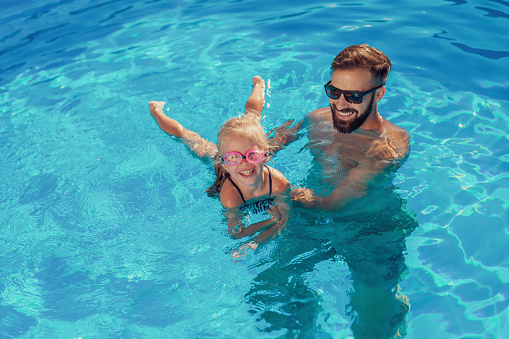 High angle view of beautiful little girl and her father having fun in the swimming pool on a hot sunny summer day, father teaching daughter to swim