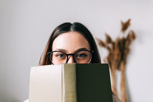 Retrato de una joven estudiante universitaria caucásica con anteojos escondido detrás de un libro y mirando a la cámara. photo