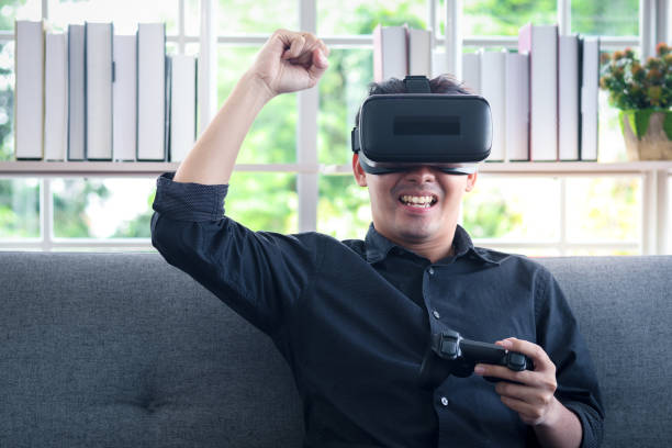 istock Asian young happy man wearing virtual reality device (VR glasses) for playing the video game while sitting on sofa in his apartment, cheerful male having fun with new trends technology, technologies of the future concept. 1301631145