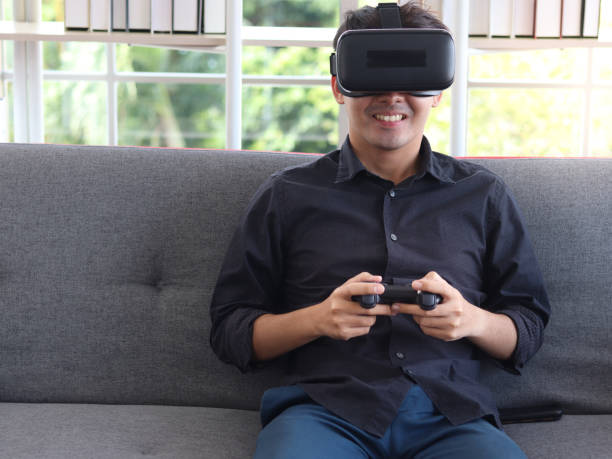 istock Asian young happy man wearing virtual reality device (VR glasses) for playing the video game while sitting on sofa in his apartment, cheerful male having fun with new trends technology, technologies of the future concept. 1301631225