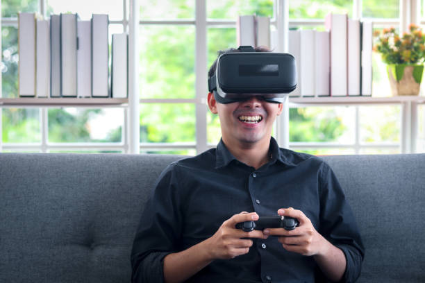 istock Asian young happy man wearing virtual reality device (VR glasses) for playing the video game while sitting on sofa in his apartment, cheerful male having fun with new trends technology, technologies of the future concept. 1301631266