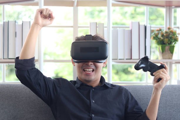 istock Asian young happy man wearing virtual reality device (VR glasses) for playing the video game while sitting on sofa in his apartment, cheerful male having fun with new trends technology, technologies of the future concept. 1301631324