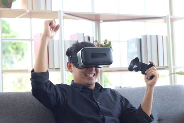 istock Asian young happy man wearing virtual reality device (VR glasses) for playing the video game while sitting on sofa in his apartment, cheerful male having fun with new trends technology, technologies of the future concept. 1301631334