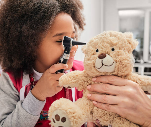 la ragazza afroamericana gioca con il suo orso giocattolo in un gioco medico, usando un otoscopio. clinica per l'udito per bambini - esame otorino foto e immagini stock