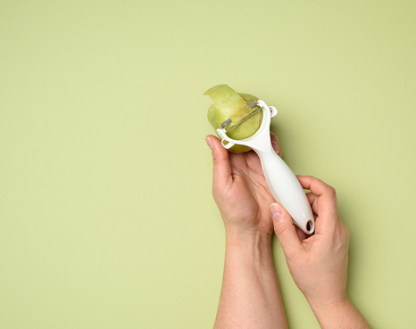 female hands hold a plastic knife for cleaning vegetables and fruits and a green apple on a green background, close up
