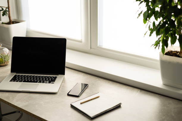 istock Still life view of office room with open laptop computer on desk with reflections by bright window, office interior. Professional workplace technology indoors. 1301648101