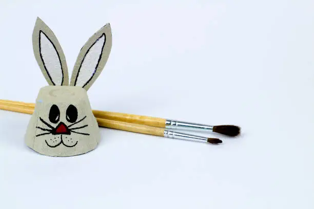 Easter bunny made of cardboard egg cup next to brushes. White background