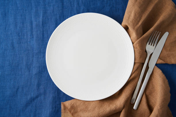 istock Clean empty white plate, fork and knife on bright blue dark linen tablecloth on table, copy space 1301651025