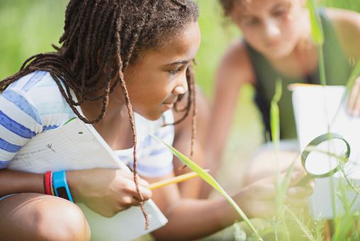 An elementary school girl of African descent is using a magnifying glass outdoors to look closer at the leaves on the ground.