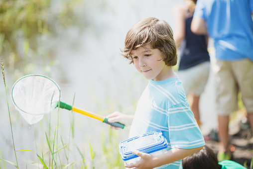 An elementary school boy of Caucasian ethnicity is outdoors exploring nature with his friends. He is holding a butterfly net.