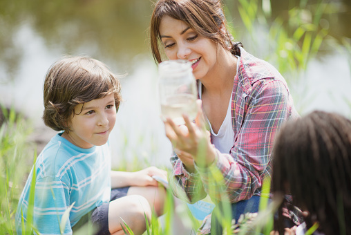 A teacher of Hispanic ethnicity is showing her student an insect that she caught in a glass jar. They are both outdoors on a warm summer day dong some exploration in nature.